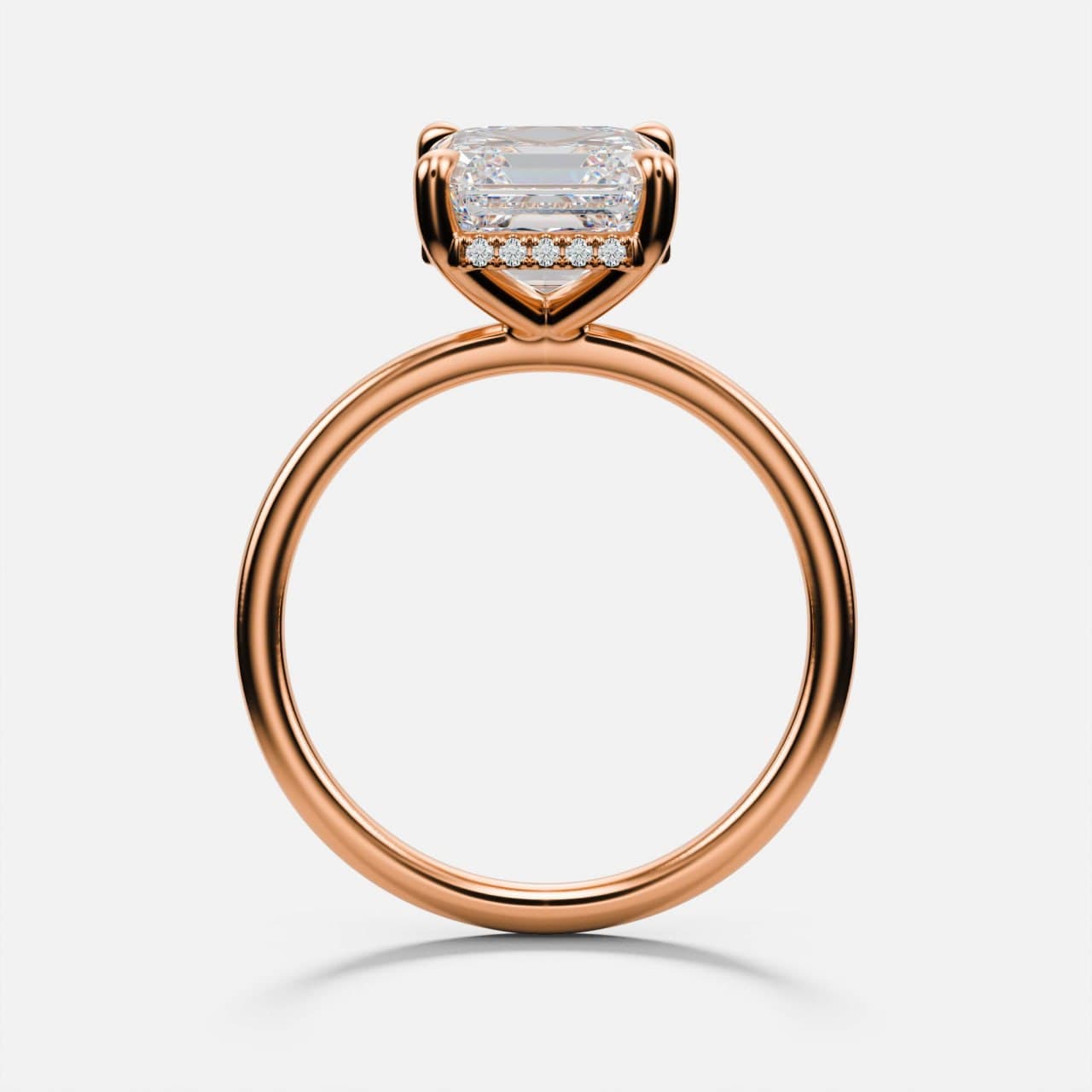 Profile view of a rose gold emerald-cut solitaire engagement ring, showing the hidden halo of pavé diamonds set beneath the centre stone and the double-claw prongs