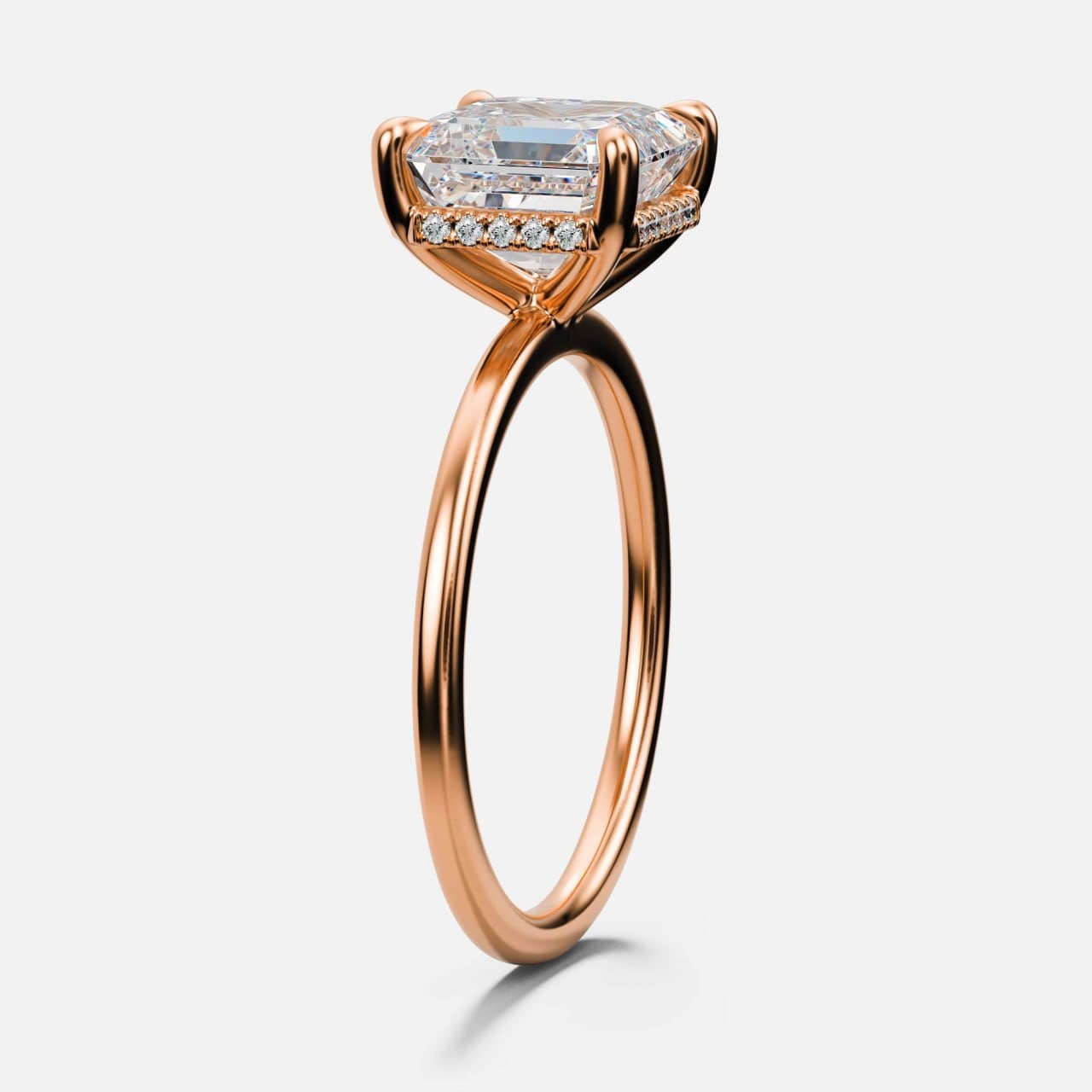 Emerald-cut diamond solitaire in rose gold on a knife-thin band, three-quarter view revealing a hidden halo of pavé diamonds beneath the centre stone — photorealistic 3D render by 4rend Studio